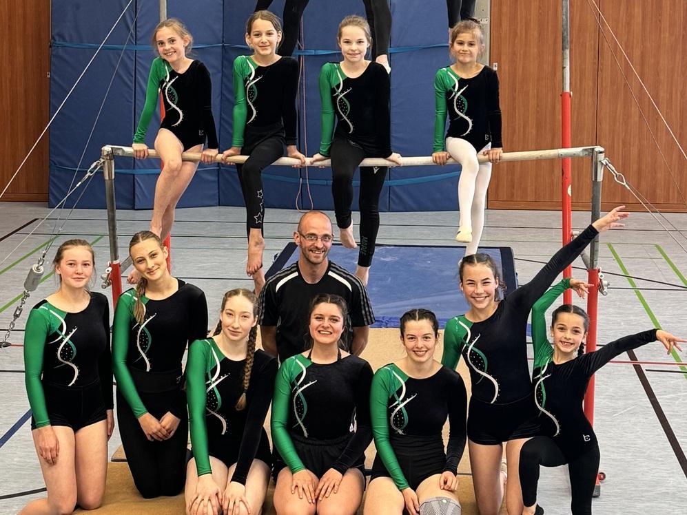 Gruppe von Turnerinnen in Schwarz-Grün, posierend auf einem Barren in einer Sporthalle mit Kletterwand.