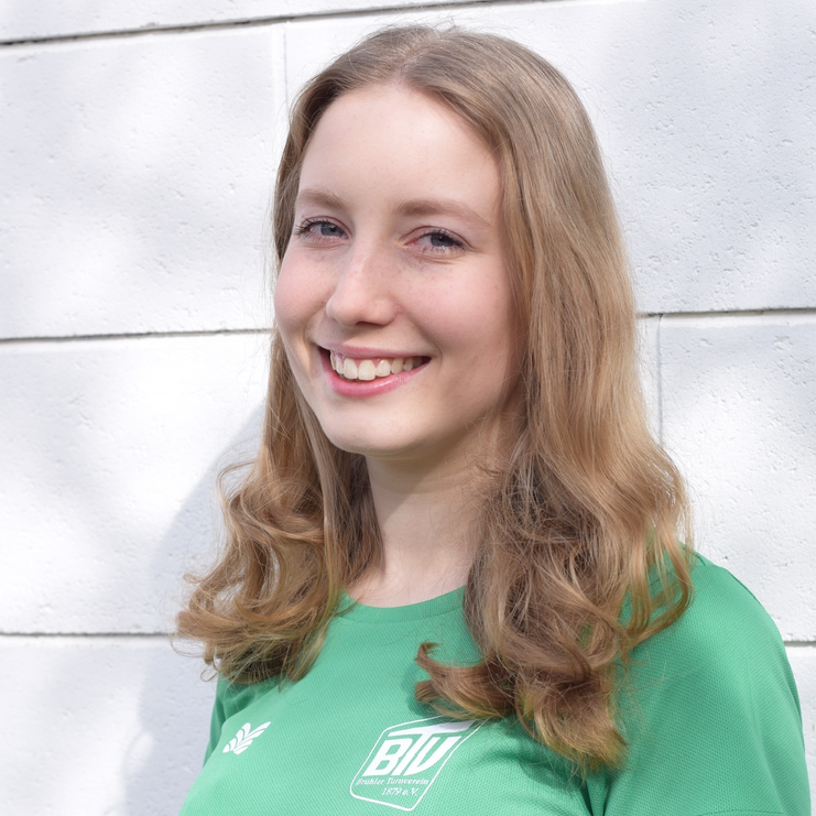 Lächelnde junge Frau mit langen, lockigen Haaren in einem grünen Shirt vor einer weißen Wand.