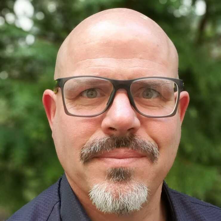 Porträt eines bärtigen Mannes mit Brille, grinsend vor grüner Hintergrundvegetation. Er trägt ein schwarzes Hemd.