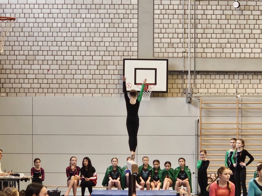 Turnerin in schwarzem Anzug balanciert auf einem Schwebebalken, während Zuschauer und Richter applaudieren.