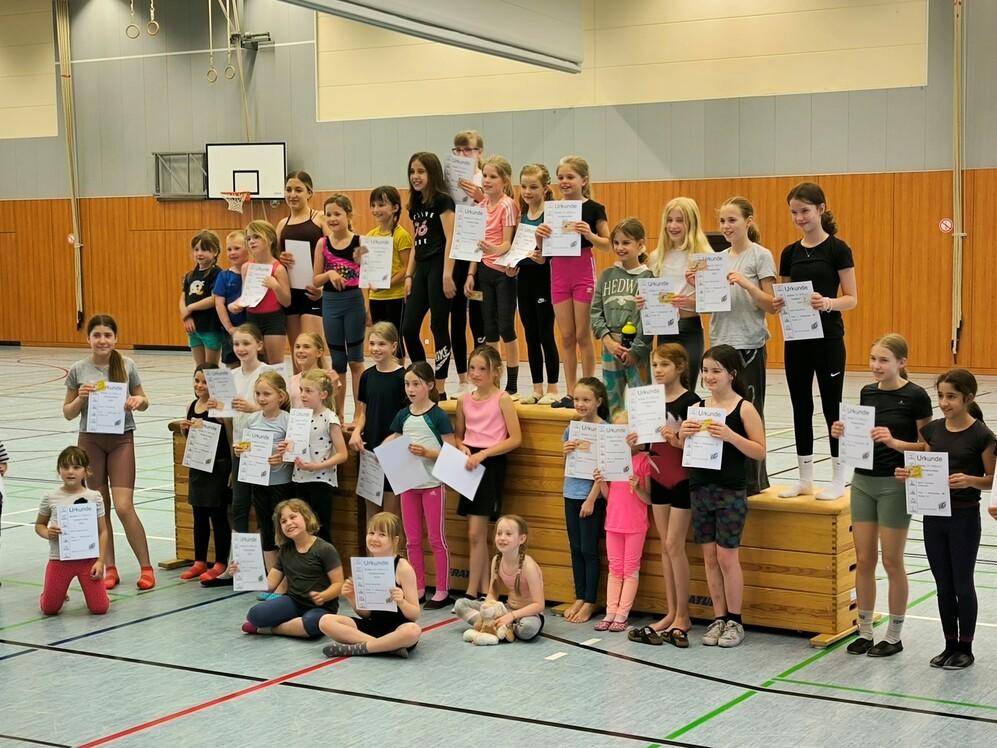Gruppe von Mädchen steht auf einer Holzplattform in einer Sporthalle und hält Urkunden in der Hand.