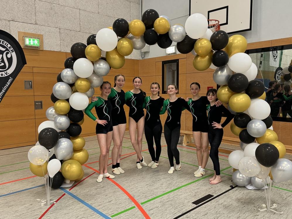 Sechs Sportlerinnen in schwarz-grünen Anzügen stehen vor einem Ballonbogen in einer Sporthalle.