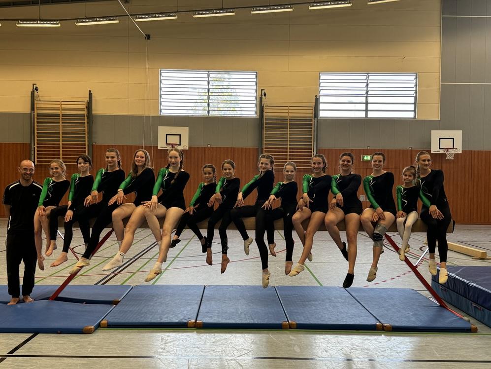 Gruppe von Turnerinnen in Schwarz-Grün sitzt auf einem Turnpferd in einer Sporthalle, Trainer steht lächelnd davor.