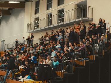 Voll besetzte Tribüne in einer Halle mit applaudierendem Publikum und Bänken im Vordergrund.