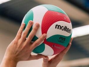 Hände heben einen bunten Volleyball mit dem Logo „molten“ in einer Sporthalle.