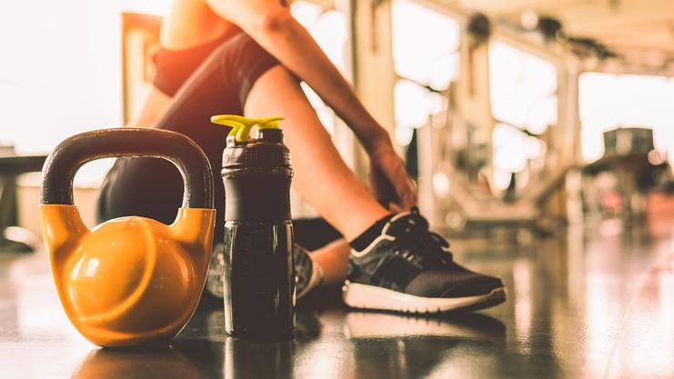 Kettlebell und Wasserflasche stehen auf dem Boden, w&auml;hrend eine Person im Fitnessstudio sitzend trainiert.