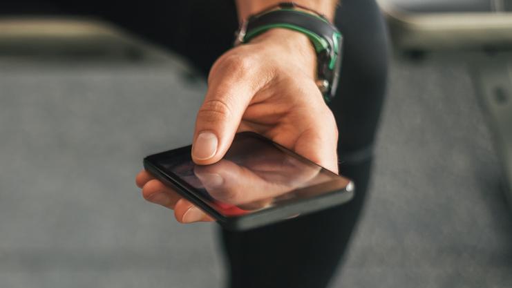 Mann sitzt auf einem Fitnessgerät und hält eine Wasserflasche in der linken Hand und ein Handy in der rechten Hand.