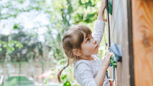 Ein Mädchen klettert an einer Holz-Kletterwand im Freien und greift nach einem Haltegriff.
