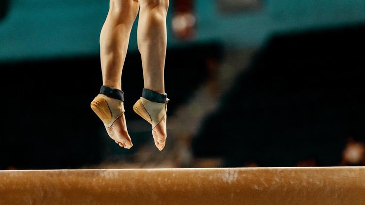 Füße eines Turnerers, die auf einem Schwebebalken balancieren, Umriss eines Wettkampfs im Hintergrund.