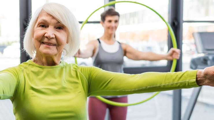 Ältere Frau in grüner Sportkleidung mit Hula-Hoop-Reifen, andere Teilnehmerin im Hintergrund. Fitnessstudio-Umgebung.