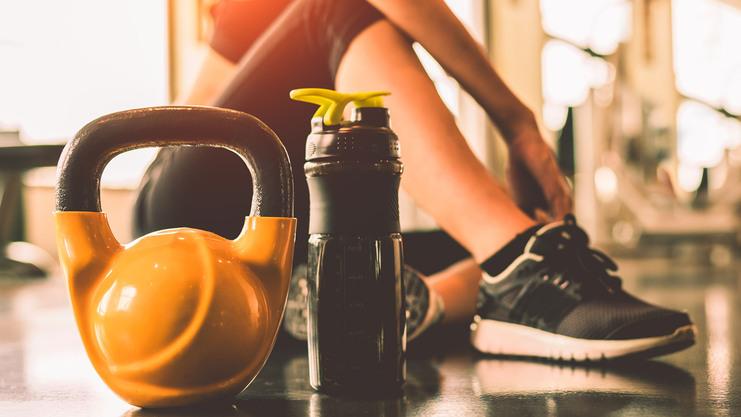 Kettlebell und Wasserflasche stehen auf dem Boden, während eine Person im Fitnessstudio sitzend trainiert.