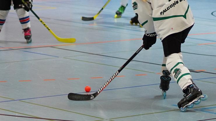 Spieler in Eishockeyausrüstung steuert einen orangen Ball mit einem Schläger auf einer Sporthalle-Bodenmarkierung.