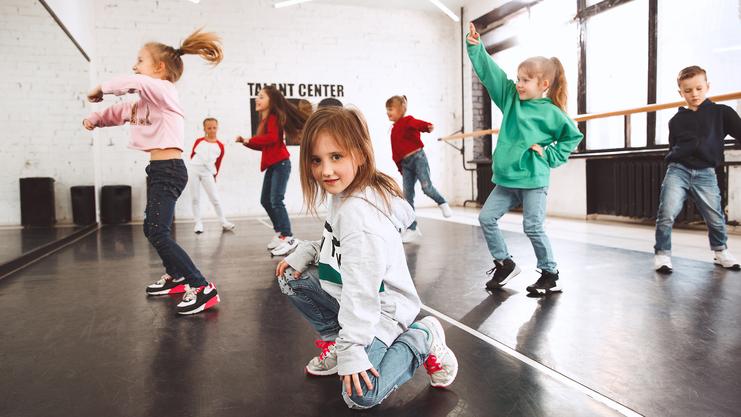Gruppe von Kindern tanzt in einem Studio, ein Mädchen hockt vorn in sportlicher Kleidung auf dem Boden.