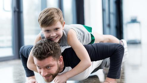 Mann macht Liegestütze, während ein Kind auf seinem Rücken sitzt und lächelt, in einem hellen Raum mit Fenster.