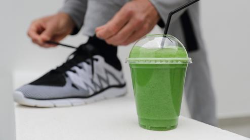 Person bindet die Schnürsenkel eines schwarzen Sneakers, neben einem grünen Smoothie in einem Becher mit Strohhalm.