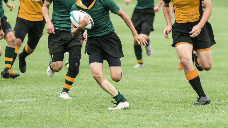 Rugbyspieler in Action, der einen Ball auf einem grünen Spielfeld trägt, umgeben von Mitspielern.