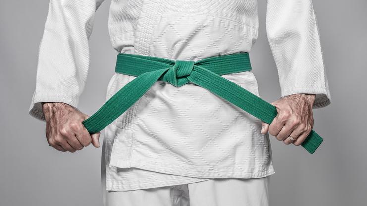 Nahaufnahme von Händen, die einen grünen Gürtel um eine weiße Gi-Jacke binden. Hintergrund grau.