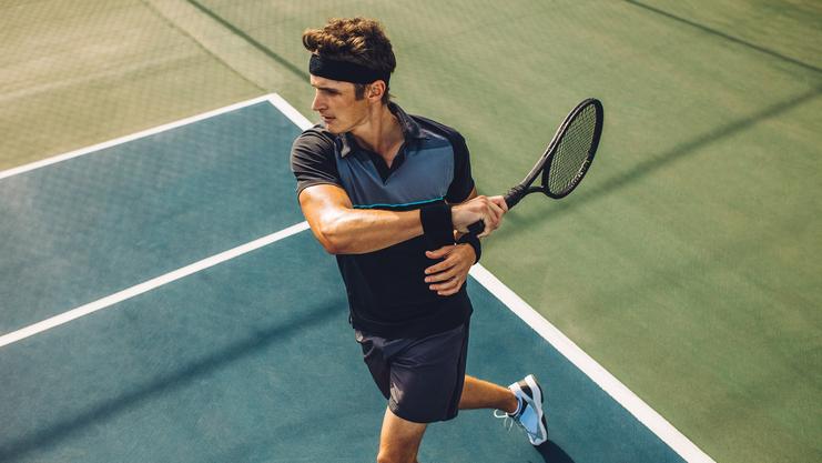 Sportler in schwarzem Shirt und schwarzem Stirnband schlägt mit einem Tennisschläger auf einem Tennisplatz.