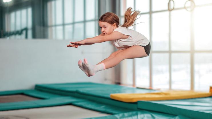 Mädchen springt mit ausgestreckten Armen über Trampolinen in einem Sportstudio mit großen Fenstern.