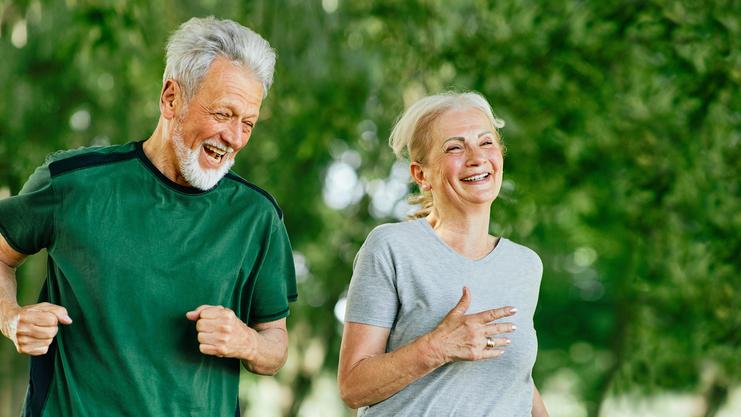 Lächelndes Seniorenpaar joggt gemeinsam auf einem gepflasterten Weg in einem grünen Park.