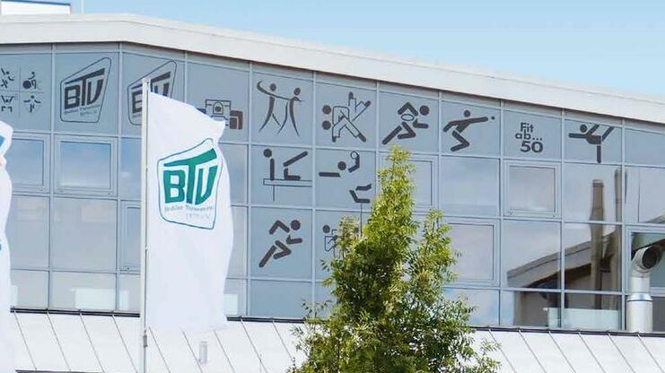 Fassade eines modernen Gebäudes mit großen Fenstern, Flaggen und sportlichen Symbolen. Grüne Pflanzen und Treppen im Vordergrund.