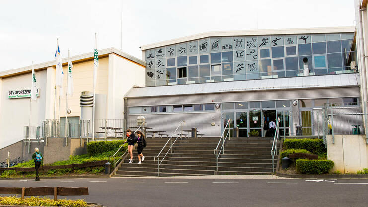 Eingang zum BTV-Sportzentrum mit Treppen, mehreren Fahnen und Radabstellplätzen in einer urbanen Umgebung.