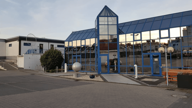 Moderne Sporteinrichtung mit gro&szlig;en Glasfronten und blauen Akzenten, umgeben von Gr&uuml;nanlagen und Parkplatz.