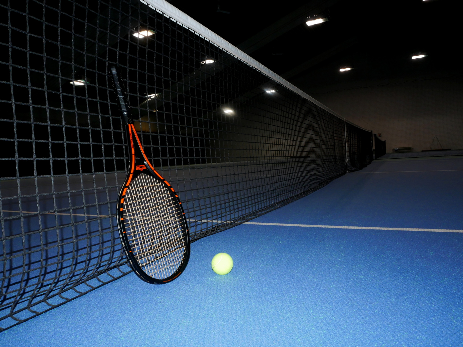 Tennisnetz mit einem Schläger und einem gelben Ball auf einem blauen Tennisplatz in einer Halle.