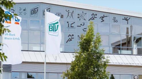 Fassade eines modernen Geb&auml;udes mit gro&szlig;en Fenstern, Flaggen und sportlichen Symbolen. Gr&uuml;ne Pflanzen und Treppen im Vordergrund.