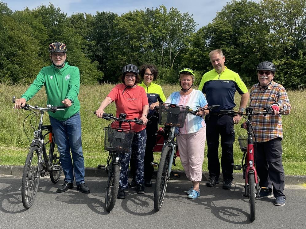 Sechs Personen stehen mit Fahrrädern an einem Weg, umgeben von einer grünen Wiese und blauem Himmel.