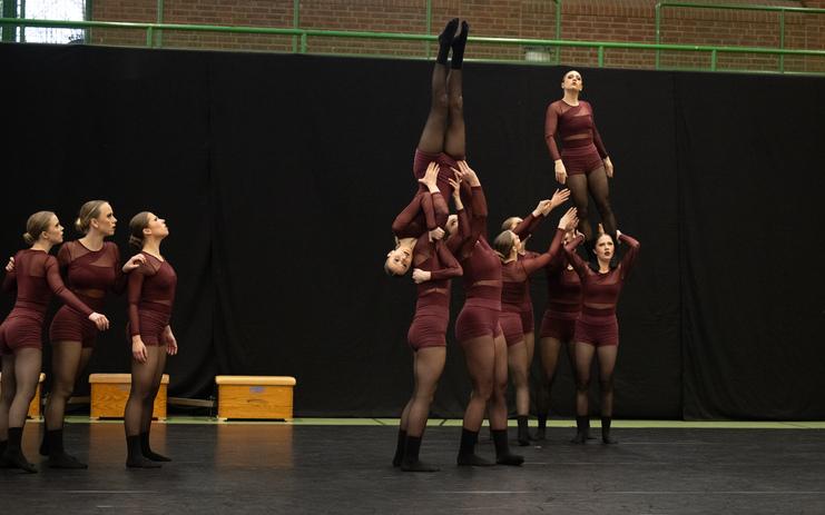 Acht Tänzerinnen in dunkelroten Outfits führen eine akrobatische Formation auf einer Sportbühne aus.