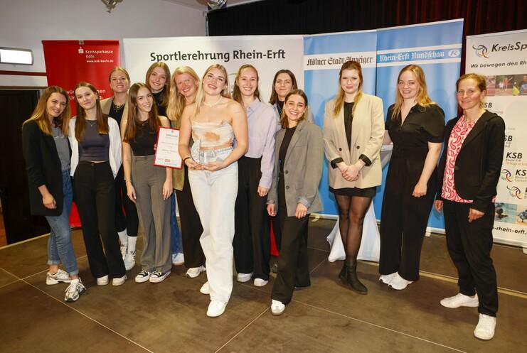 Gruppe von 12 Frauen bei einer Sportlerehrung, eine hält eine Urkunde in der Hand. Bunte Banner im Hintergrund.