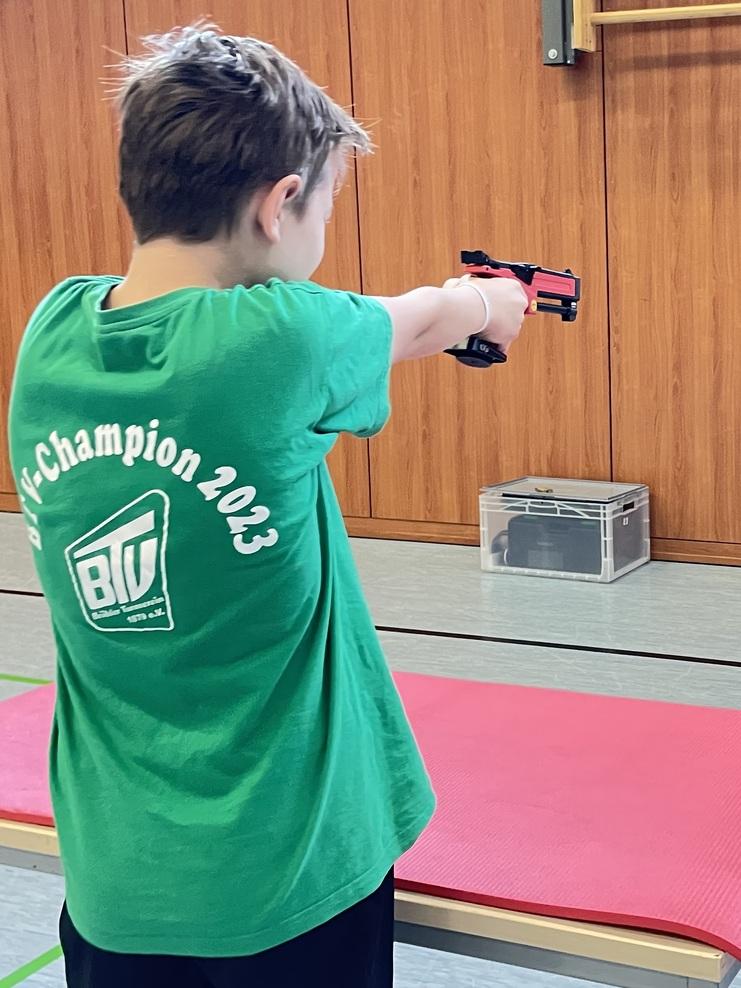 Ein Kind in grünem T-Shirt zielt mit einer Sportpistole auf einen Zielplatz in einer Sporthalle.