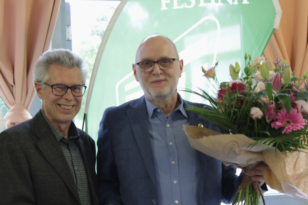 Zwei Männer lächeln, der rechte hält einen Blumenstrauß in der Hand, im Hintergrund eine grüne Bannerfläche.