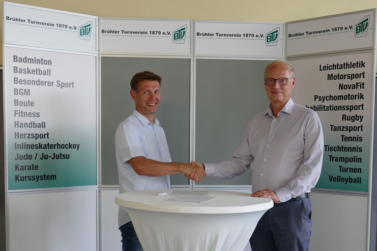Zwei M&auml;nner sch&uuml;tteln sich die H&auml;nde vor einem Banner des Br&uuml;hler Turnvereins 1879 e.V. und einem Stehtisch.