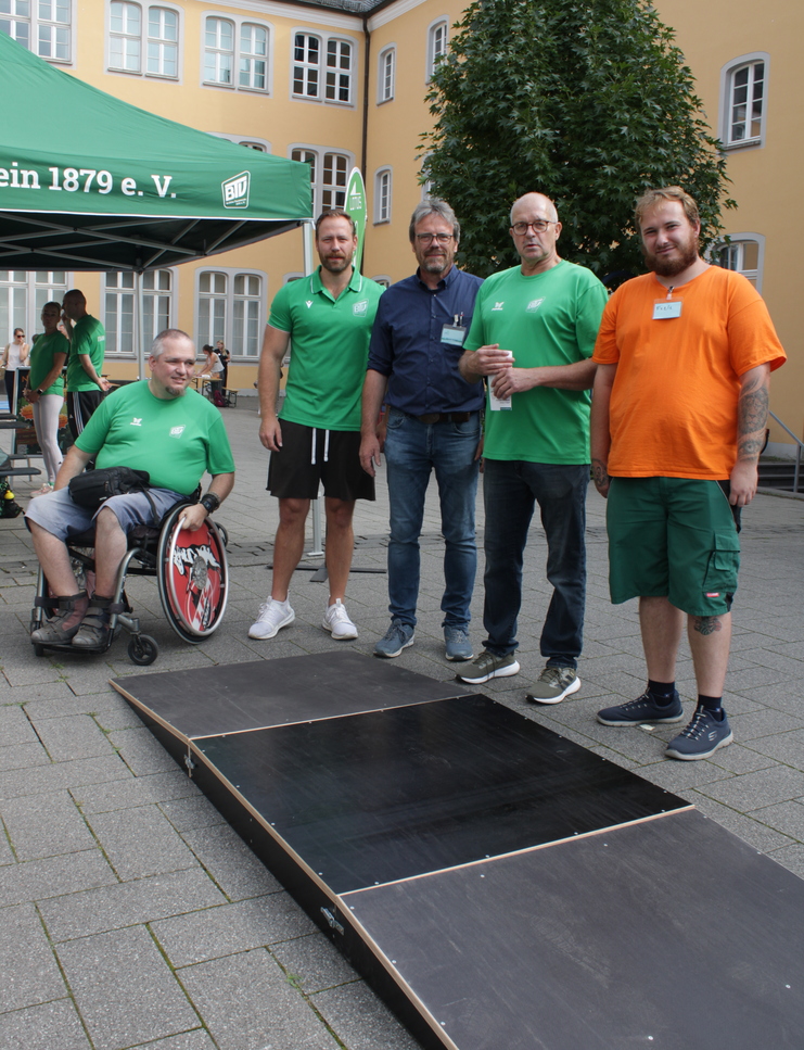 Fünf Männer, darunter ein Rollstuhlfahrer, stehen vor einer Rampe und einem Zelt des Vereins 1879 e.V. in einem Innenhof.