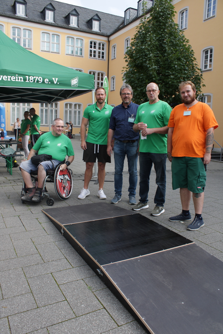 Fünf Männer, darunter ein Rollstuhlfahrer, stehen vor einer Rampe und einem Zelt des Vereins 1879 e.V. in einem Innenhof.