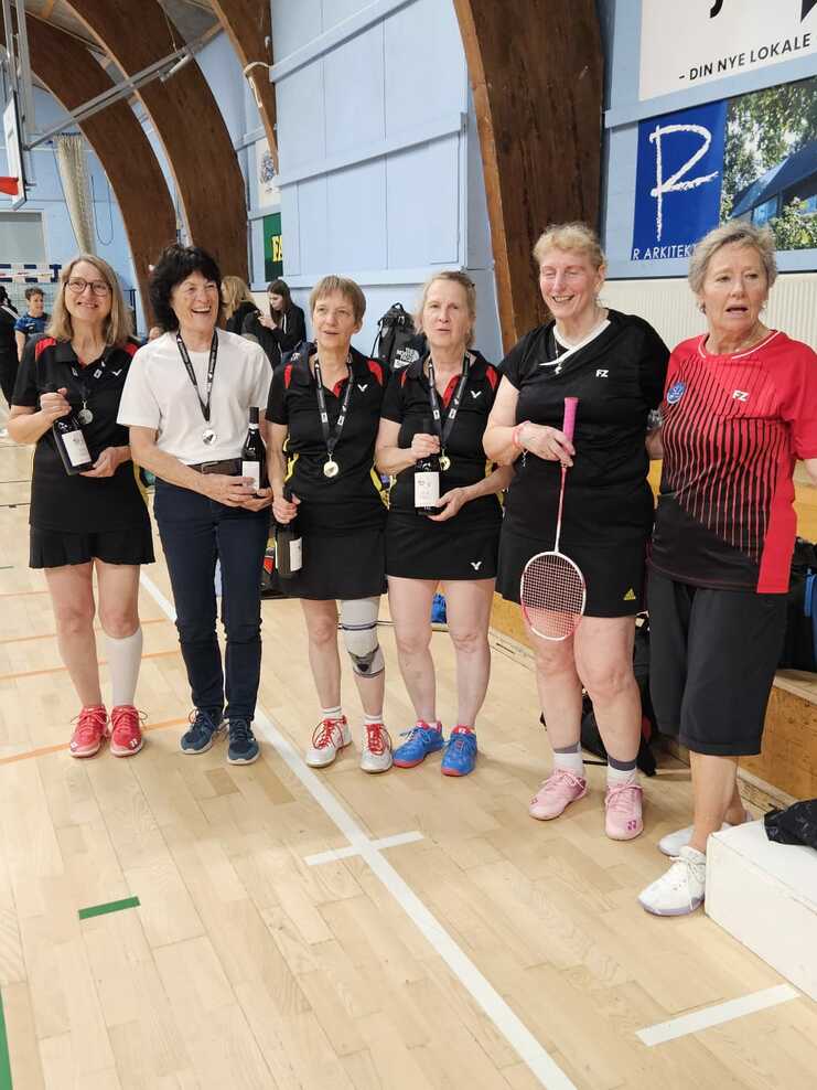 Gruppe von sechs Badminton-Spielerinnen mit Medaillen in Sportkleidung in einer Halle. Hintergründe zeigen sportliche Aktivitäten.