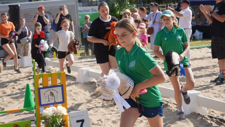 Mädchen in grünen Shirts laufen im Sand mit Plüschpferden zu einem Wettkampf, Zuschauer applaudieren im Hintergrund.