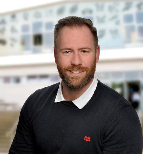 Porträt eines Mannes mit Bart, der in einem schwarzen Pullover mit weißem Kragen vor einem unscharfen Gebäude lächelt.