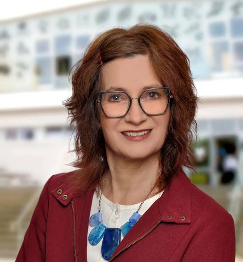 Porträt einer lächelnden Frau mit Brille, braunen Haaren und blauer Halskette, vor einem unscharfen Hintergrund.