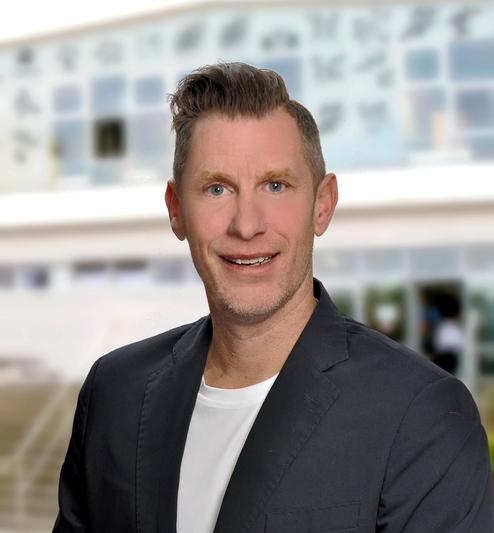 Porträt eines Mannes mit kurzem Haar, in dunklem Blazer und weißem Shirt, vor einem verschwommenen Gebäude.