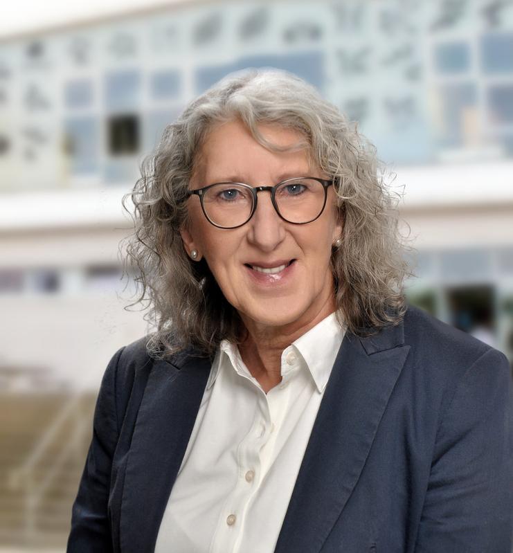 Portrait einer lächelnden Frau mit grauen, lockigen Haaren, Brille und Blazer, stehend vor unscharfem Hintergrund.