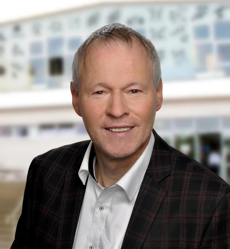 Portrait eines lächelnden Mannes in einem karierten Sakko vor unscharfem Hintergrund eines Gebäudes.