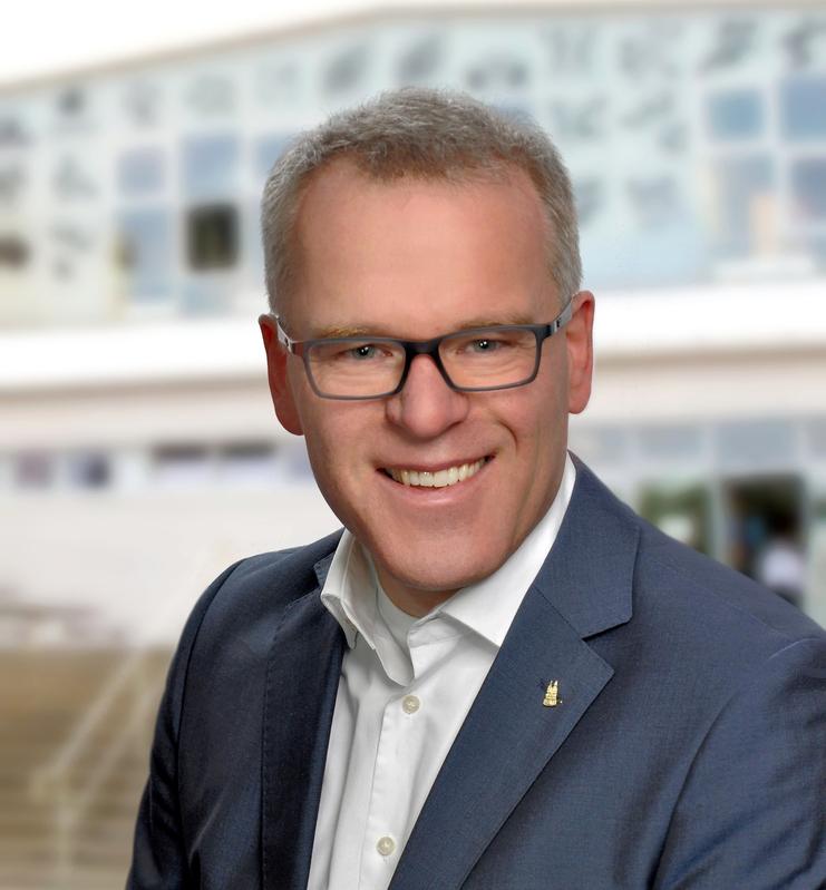 Porträt eines lächelnden Mannes mit Brille in Anzug, stehender Kopf, unscharfer Hintergrund mit moderner Architektur.