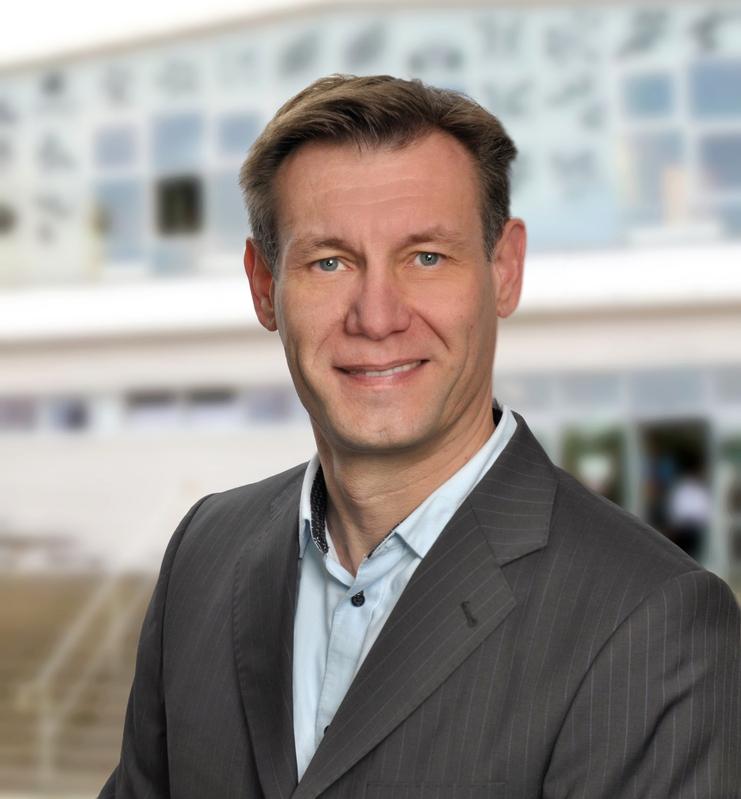 Porträt eines Mannes in einem Anzug mit grünem Hemd, lächelnd vor verschwommenem modernen Hintergrund.