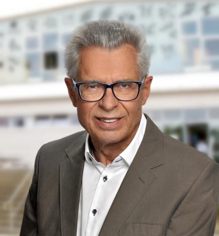 Porträt eines Mannes mit grauen Haaren und Brille, in einem hellen Sakko vor einem modernen Gebäude.